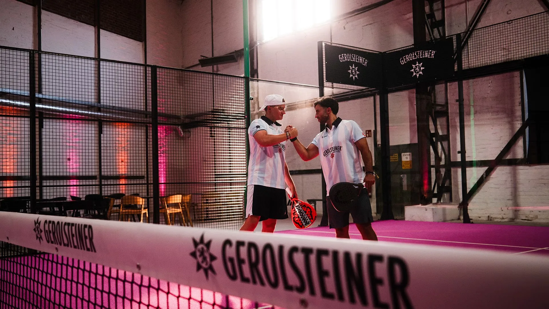 Two padel players bumping fists on a magenta indoor court, Gerolsteiner on the net